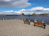 A serene view of the Jacques-Cartier Marina in Gatineau, Québec, with a wide river bordered by a clear sky with fluffy clouds. Two empty benches face the water, inviting a moment of relaxation. The background features a bridge spanning the river and a cityscape with various buildings.